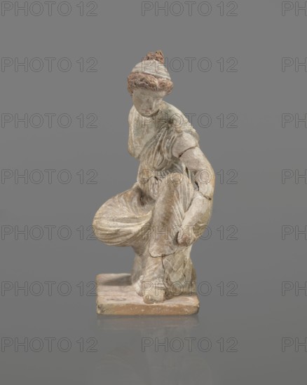 Terracotta figurine of a girl throwing dice. Figurine of a crouching girl throwing dice: weight of the body rests on the left leg bent at a right angle. Dressed in a long, finely pleated, sleeveless chiton. Hair tied in a bun and bound with a wide headband kredemnos. Earring visible in the left ear. Entire figurine rests on a rectangular base. Preservation: missing the right arm; glued in several places head; left arm from 2 fragments; robe; base from 3 fragments. Part of the base filled with clay and plaster. Date: 4th–3rd century BC. Place of origin: Tanagra Greece. Material/type: terracotta mold figurine, Tanagrian style. Dimensions: height 13 cm, width 9 cm, depth 4.5 cm., A small carved figure of a person seated on a rectangular base, shown against a plain gray background; the figure wears draped clothing with visible folds and has hair pulled back into a bun, with traces of reddish-brown and pale beige pigments across the surface and areas of worn white and gray; the sculpture sh...