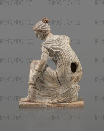 Terracotta figurine of a girl throwing dice. Figurine of a crouching girl throwing dice: weight of the body rests on the left leg, bent at a right angle. She wears a long, finely pleated, sleeveless chiton. Hair tied in a bun with a wide headband kredemnos. Earring visible in left ear. Entire figurine rests on a rectangular base. Preservation: missing right arm; glued in several places head, left arm from 2 fragments, robe, base from 3 fragments. Part of the base filled with clay and plaster. 4th–3rd century BC, Tanagra Greece. Terracotta; height 13 cm, width 9 cm, depth 4.5 cm., A weathered sculpted figure in muted beige and gray tones sits on a rectangular base, shown in profile leaning forward with a draped garment that has carved folds and a textured back; the figure’s hair is styled in a coiled bun with subtle reddish-brown accents, the skin areas show pale beige with worn spots, and there is a dark circular hole visible on the figure’s side; cracks and repairs are visible across ...