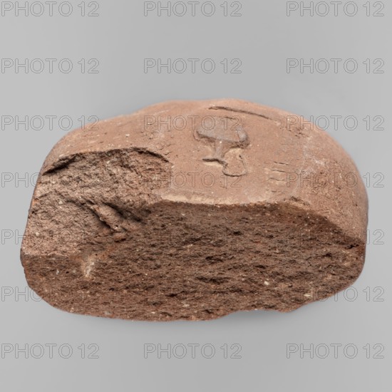 Fragment of a transport amphora vice with a stamp depicting an oinochoe and an incomplete Greek inscription. Fragment of an amphora vice with a rectangular stamp containing an inscription in two rows and between them a depiction of an oinochoe in a horizontal position. End of the 4th–beginning of the 3rd century BC. Found at Myrmekion; origin Thasos. Stamp; vice. Height 5.1 cm, width 10.8 cm., A rough, irregularly shaped reddish-brown clay fragment rests against a plain light gray background; the fragment shows a mix of coarse and smoother textures with a rougher, grainy underside and a more compact upper surface that includes an incised shallow curved impression and a rounded raised mark, and there are small flecks of lighter and darker tones within the reddish-brown body.