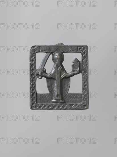 Icon: St. Nicholas Mozajski. Orthodox workshop; Nicholas of Myra St.; ca. 270–345/352. The icon shows St. Nicholas holding a model of the city in his left hand and raising a sword in his right. The background is cut out, with an identifying inscription on the upper frame. The relief of the cast is schematic with extensive ornamentation; proportions are elongated, head and hands enlarged relative to the body. 1st half of the 18th century, Russia; icon. Height 6.4 cm, width 5.9 cm., A small dark gray metal plaque with a square frame featuring decorative interlaced patterns along its borders and a textured background, centered on a light gray background. Inside the frame is a raised figure with a rounded head and draped robe, standing on a small pedestal; the figure holds a curved object in one hand and a block-like object in the other. The metal surface shows slight variations in darker and lighter gray tones and subtle reflective highlights.