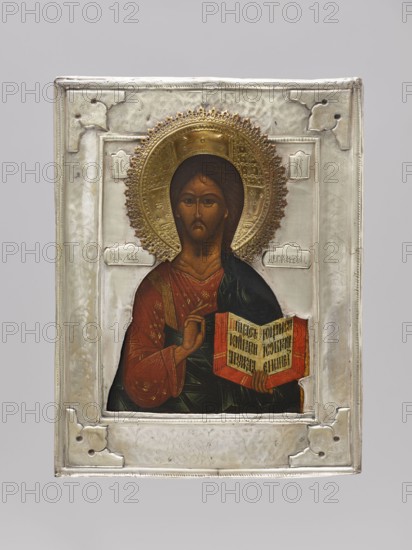 Icon in a cover: Christ Pantocrator. The icon represents a traditional, half-length variant of Christ Pantocrator referring to designs dating to the 16th century. The Savior's face is elongated, ascetic, with a light brown complexion brightened by tones; lower eyelids, forehead wrinkles, and neck folds are clearly marked. Christ wears a red chiton decorated with fine gold ornaments, an ochre sash covered with assyst, and a navy blue himation with rather small, pointed folds. The inscription on the book is made with a decorative ribbon. Around Christ's head is a cross halo with the letters OOH. On both sides of the figure are inscriptions: the Christological monogram and Lord Almighty. The entire field, the icon background, and the halo are covered with a brass cover with an applied wreath. 18th century, Russia; icon with cover; height 31 cm, width 24 cm., An icon painting of a figure with a golden halo set within a silver frame, showing a person with brown hair and a brown beard wearin...