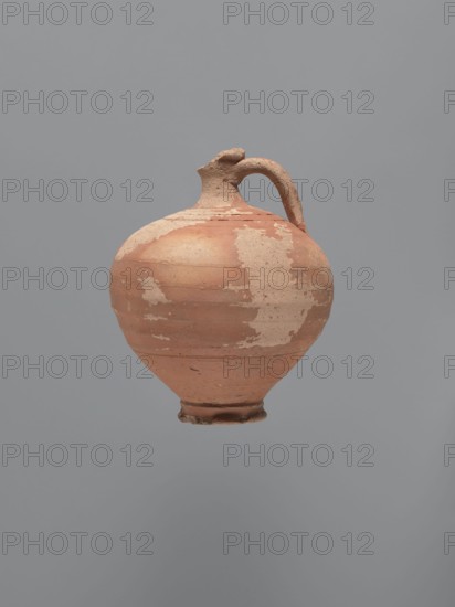 Jug, unknown Aswan workshop; type: jug; materials: kaolin ceramic mass Gempeler type IA with exterior red-brown engobe and interior beige-pink; form: rounded rim, narrow upward-flaring neck with ring-shaped flange, spherical body with a large ribbon-shaped handle attached to lower body and mid-neck, ring-shaped foot; traces of turning slightly smoothed; style/technique: Egyptian sigillata/Coptic pottery/tableware; date: 5th–6th century; find location: Edfu; dimensions: height 14.1 cm; lip diameter 2.4 cm; neck diameter 1.8 cm; body diameter 11.4 cm; base diameter 4.6 cm., A rounded ceramic jug with a single curved handle is shown against a plain gray background, the jug exhibiting a weathered surface with patches of lighter and darker terracotta tones and areas where the outer layer has worn away, revealing a paler, sandy color; the jug has a narrow neck, a small spout, and a slightly flared foot, and its surface shows irregular horizontal lines and rough texture in shades of reddish-b...