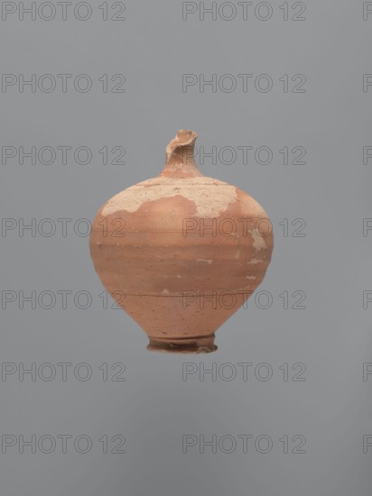 Jug, unknown Aswan workshop. Jug with a rounded rim, a narrow and upwardly flaring neck with a ring-shaped flange. Spherical body with a large ribbon-shaped handle glued to the lower part of the body and the middle part of the neck. Ring-shaped foot. Traces of turning slightly smoothed. Exterior covered with red-brown engobe, interior beige-pink, even. Kaolin ceramic mass Gempeler type IA. Egyptian sigillata, Egyptian pottery, Coptic vessels, tableware. 5th–6th century. Edfu find location. Vessel, jug; height 14.1 cm; lip diameter 2.4 cm; neck diameter 1.8 cm; body diameter 11.4 cm; base diameter 4.6 cm., A rounded clay vessel with a narrow base and a short, irregular neck suspended against a plain background; the vessel shows worn and flaked surfaces with patches of lighter and darker terracotta tones, a few areas of pale beige residue, and a generally matte, textured appearance in shades of reddish-brown, beige, and muted orange against a solid gray backdrop.
