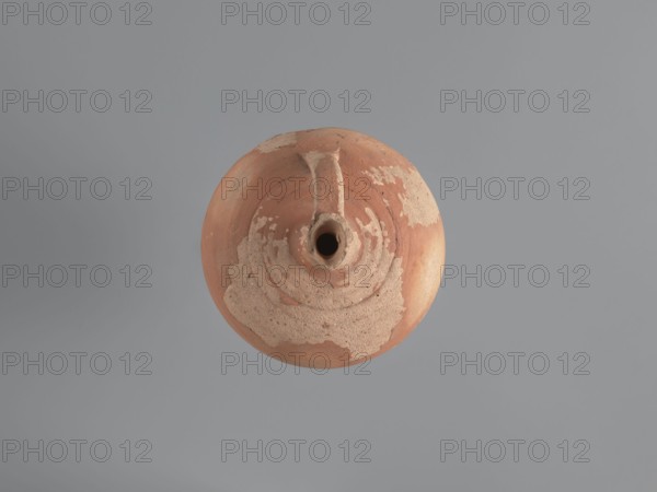 Jug, unknown Aswan workshop. Jug with a rounded rim, a narrow and upwardly flaring neck with a ring-shaped flange. Spherical body with a large ribbon-shaped handle attached to the lower part of the body and the middle part of the neck. Ring-shaped foot. Traces of turning are slightly smoothed. Exterior covered with red-brown engobe; interior beige-pink, even. Kaolin ceramic mass Gempeler type IA. Egyptian sigillata/Coptic pottery, tableware, kaolin clay. 5th–6th century. Findspot: Edfu. Vessel jug. Height 14.1 cm; lip diameter 2.4 cm; neck diameter 1.8 cm; body diameter 11.4 cm; base diameter 4.6 cm., A round, worn clay vessel sits centered against a smooth grey background, showing a top view with a small central opening and a short spout. The vessel displays a mix of reddish-brown and beige tones, with patches of lighter beige where the surface appears chipped or eroded, and circular ridges encircling the opening. The grey background appears uniform with subtle darker grey shading tow...