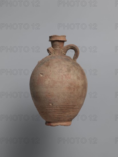 Bottle, unknown Egyptian workshop; painted Coptic pottery vessel bottle, marl clay with limestone inclusions, possibly sealed/repaired with bitumen. Rounded, falling rim; concavely profiled, almost cylindrical neck flowing into rounded shoulders decorated with fine ribs. Two handles one fragmentary attached at lower neck and upper shoulders. Ovoid body with grooved lower body turning traces and ring-shaped foot. Outer surface brown and smoothed. Faint black-painted decoration on shoulders: two arcades of parallel arches separated by a row of dots, arranged symmetrically one arcade between the handles. Irregular concentration of black substance at the highest bulge of the body below the damaged handle, likely bitumen. 6th century; from Edfu. Height 22.7 cm; spout diameter 4.3 cm; neck diameter 3.0 cm; belly diameter 14.9 cm; bottom diameter 7.2 cm., A weathered earthenware vessel with a rounded body and a narrow neck topped by a flared rim, showing rough textured surface and small chips...