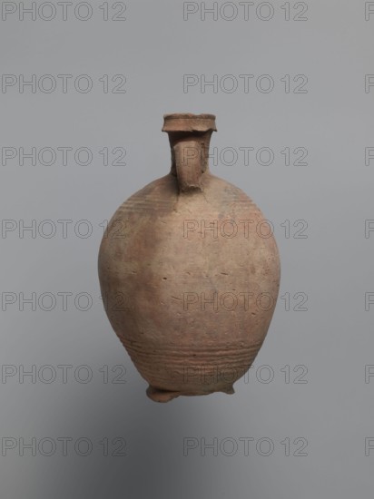 Bottle, unknown Egyptian workshop; painted Coptic pottery bottle, marl clay with limestone inclusions, possibly sealed/repaired with bitumen. Rounded, falling rim, concavely profiled; almost cylindrical neck flowing into rounded shoulders covered with fine ribs. Two handles one fragmentary attached to lower neck and upper shoulders. Ovoid body with lower body grooved turning traces and ring-shaped foot. Outer surface brown and smoothed. Faint black painted decoration on shoulders: two symmetrical arcades of parallel arches separated by a row of dots one arcade between the handles. Irregular concentration of black substance at highest bulge below damaged handle likely bitumen. 6th century, Edfu; vessel bottle. Height 22.7 cm; spout diameter 4.3 cm; neck diameter 3.0 cm; belly diameter 14.9 cm; bottom diameter 7.2 cm., An earthenware vessel with a rounded body and a narrow neck topped by a short flared rim, showing a rough, aged surface with small pits and uneven texture; the vessel has ...