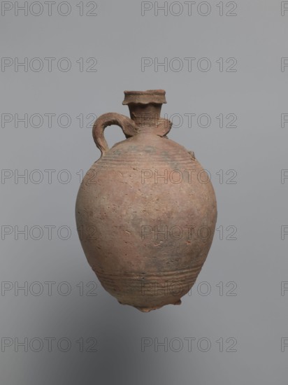 Bottle, unknown Egyptian workshop; painted Coptic pottery vessel, 6th century, from Edfu. Bottle with a rounded, falling rim and concavely profiled neck. The almost cylindrical neck flows into rounded shoulders covered with fine ribs. Two handles one fragmentarily preserved are attached to the lower part of the neck and upper part of the shoulders. The ovoid body shows grooves on its lowest part traces of turning and rests on a ring-shaped foot. Outer surface brown and burnished. Made of A4 marl clay with limestone inclusions. Shoulders bear faint black-painted decoration: two arcades of parallel arches separated by a row of dots, arranged symmetrically one arcade between the handles. At the highest bulge of the body, below the damaged handle, there is an irregular concentration of a black substance, likely bitumen used to seal or repair the wall. Function: vessel/bottle. Dimensions: height 22.7 cm; spout diameter 4.3 cm; neck diameter 3.0 cm; belly diameter 14.9 cm; bottom diameter 7....