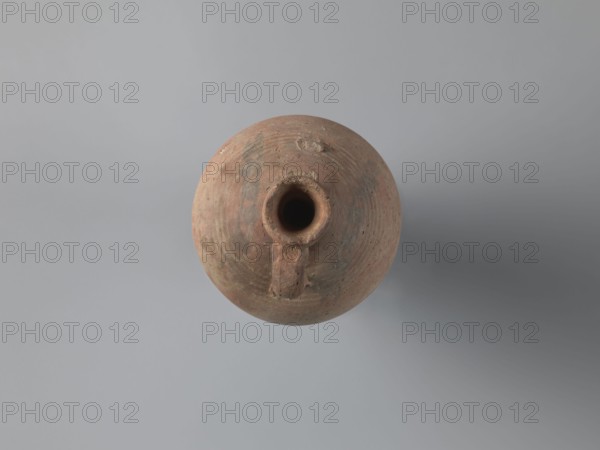 Bottle, unknown Egyptian workshop; painted Coptic pottery, marl clay with limestone inclusions, with bitumen residue. Bottle with a rounded, falling rim, concavely profiled. Almost cylindrical neck flows into rounded shoulders covered with fine ribs. Two handles one fragmentary are attached to the lower neck and upper shoulders. Ovoid body with lowest part grooved turning traces; ring-shaped foot. Outer surface brown and smoothed. Faint black-painted decoration on the shoulders: two arcades of parallel arches separated by a row of dots, arranged symmetrically one between the handles. Irregular concentration of black substance at the highest bulge of the body below a damaged handle likely bitumen used to seal or repair. 6th century, Edfu. Vessel bottle. Height 22.7 cm; spout diameter 4.3 cm; neck diameter 3.0 cm; belly diameter 14.9 cm; bottom diameter 7.2 cm., A round, weathered clay vessel viewed from above with a central opening and a short spout-like projection, showing a rough, tex...