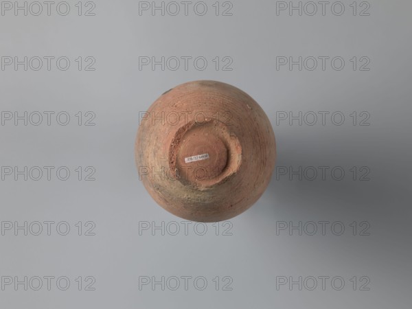 Bottle, unknown Egyptian workshop. Bottle with a rounded and falling rim, concavely profiled. The almost cylindrical neck flows into rounded shoulders covered with fine ribs. Two handles, one fragmentarily preserved, are glued to the lowest part of the neck and the lower part of the shoulders. The ovoid body is grooved on its lowest part traces of turning; the bottom is a ring-shaped foot. Outer surface brown and burnished. Made of A4 marl clay with limestone inclusions. On the shoulders is faint painted decoration in black: two arcades composed of parallel arches separated by a row of dots, arranged symmetrically, one between the handles. At the highest bulge of the body, below the damaged handle, is an irregular concentration of a black substance, probably bitumen used to seal or repair the wall. 6th century, Edfu. Vessel bottle. Height 22.7 cm; spout diameter 4.3 cm; neck diameter 3.0 cm; belly diameter 14.9 cm; bottom diameter 7.2 cm., A round, weathered clay pot viewed from above,...