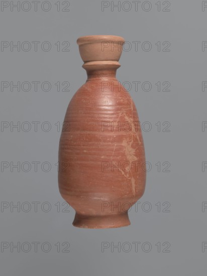 Bottle, unknown Aswan workshop; baggy-shaped bottle without preserved spout. The neck widens toward the top with a circumferential bulge and another at its base. Sloping shoulders; belly is baggy, flowing into the base on a low foot. Pink kaolin ceramic mass Gempeler IA. Partially smoothed turning marks visible on the surface. Outer walls covered with polished red engobe. Egyptian sigillata/Coptic pottery; Egyptian imports in Nubia. 2nd half 5th–1st half 6th century; find from Edfu. Vessel bottle. Height 20.6 cm; diameter 3.4 cm neck, 11.3 cm body, 7.7 cm bottom., A single pottery vessel centered against a solid gray background, with an elongated rounded body tapering to a narrow neck and flared rim. The vessel shows a smooth, slightly worn surface with visible horizontal turn lines and a few irregular pale streaks and blemishes on its body. The colors visible are terracotta, light brown, pale beige, and gray from the background. The rim and base appear slightly lighter in color than t...