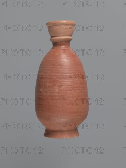 Bottle, unknown Aswan workshop; baggy-shaped bottle without preserved spout. Neck widens toward the top with a circumferential bulge and another at its base; sloping shoulders; baggy belly flowing into the base on a low foot. Pink kaolin ceramic mass Gempeler IA. Partially smoothed turning marks visible on the surface. Outer walls covered with polished red engobe. Egyptian sigillata/Aswan pottery; Coptic pottery/vessels. 2nd half 5th–1st half 6th century. Findspot: Edfu. Vessel bottle. Height 20.6 cm; diameter 3.4 cm neck, 11.3 cm body, 7.7 cm bottom., A single tall, rounded ceramic vessel with a narrow neck and flared lip, shown against a plain gray background; the vessel has a smooth, slightly textured surface in shades of reddish-brown and terracotta with some lighter beige areas around the rim and base, and faint horizontal striations visible on its body.