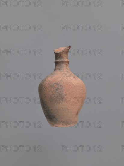 Jug, unknown Aswan workshop; jugs; tableware; Egyptian sigillata; Aswan pottery; Egyptian pottery; Coptic pottery; kaolin clay; defective firing. Jug with a rounded and outward-turning rim, a cylindrical neck tapering at the base and provided with circumferential rollers midway up and at the base. Sloping shoulders merge into an ovoid belly. A preserved trace of a vise indicates it connected the middle part of the neck with the upper part of the shoulders. Flat bottom, curved inward. Pink kaolin ceramic body Gempler IA. Surfaces eroded, but remnants of a yellow engobe survive in the bottom section. Below the vice attachment point, on the left side near the bottom, a dent in the wall is visible, probably the result of a firing error. 5th–7th century. Findspot: Edfu. Vessel jug. Height 15.7+ cm preserved; rim diameter 5.0 cm; neck diameter 2.4 cm; body diameter 9.7 cm; bottom diameter 4.0 cm., A single weathered clay vessel with a rounded body and a narrow neck is centered against a plai...