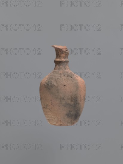 Jug, unknown Aswan workshop. Jug with a rounded outward-turning rim, cylindrical neck, tapering at the base and provided with circumferential rollers midway up and at the base. Sloping shoulders merge into an ovoid belly. A preserved trace of a vise indicates it connected the middle part of the neck with the upper part of the shoulders. Flat, inward-curving bottom. Pink kaolin ceramic body Gempler IA. Surfaces eroded, but remnants of a yellow engobe survive in the bottom section. Below the vice attachment point, on the left side near the bottom, a dent in the wall is visible, probably from a firing error. Egyptian sigillata/Coptic pottery, kaolin clay; defective firing. 5th–7th century. Find location: Edfu. Vessel type: jug. Height 15.7+ cm preserved; rim diameter 5.0 cm; neck diameter 2.4 cm; body diameter 9.7 cm; bottom diameter 4.0 cm., A worn, roughly textured clay vessel with an irregular narrow neck and chipped rim floats against a plain gray background, showing colors of reddish...
