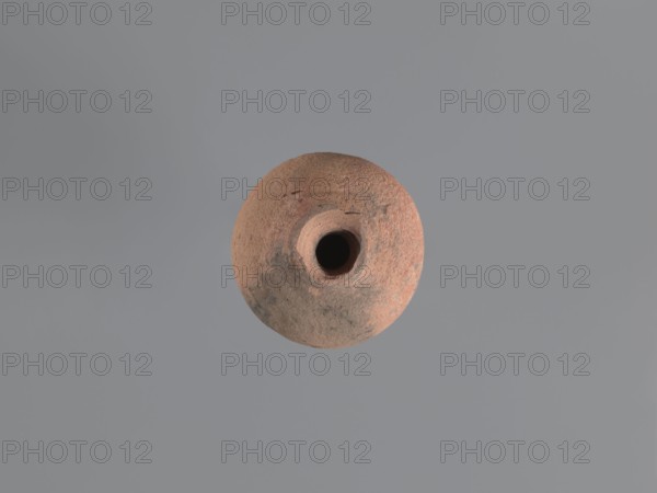 Jug, unknown Aswan workshop; jugs, tableware, Egyptian sigillata, Aswan pottery, Egyptian pottery, Coptic pottery; kaolin clay; defective firing. Jug with a rounded outward-turning rim, cylindrical neck tapering to the base, circumferential rollers midway and at the base. Sloping shoulders merge into an ovoid belly. Preserved trace of a vise indicating connection of the middle neck to the upper shoulders. Flat bottom, curved inward. Pink kaolin ceramic body Gempler IA. Surfaces eroded with remnants of a yellow engobe in the bottom section. Dent in the wall below the vice attachment point on the left side near the bottom, likely from a firing error. 5th–7th century, Edfu find location. Vessel, jug. Height 15.7+ cm preserved; rim diameter 5.0 cm; neck diameter 2.4 cm; body diameter 9.7 cm; bottom diameter 4.0 cm., A round, textured object with a central circular opening sits against a smooth gray background; the object shows earthy tones of reddish-brown, terracotta, and muted gray with ...