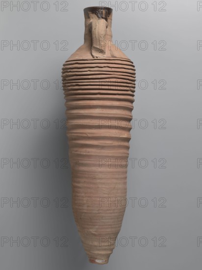 Amphora, unknown Egyptian workshop; amphorae, Egyptian amphorae, Egyptian ceramics, storage vessels, Coptic vessels, amphorae; Nile clay. Wine transport vessel with a spindle-cylindrical, long body. Steep arms, cylindrical neck ending with a straight rim. Cylindrical viscæ, oval in cross-section, are attached to the lower part of the arms and to the middle part of the neck. The entire body has grooves of varying density and size; the densest are in the lower and upper parts. No cylindrical foot. Brown, alluvial clay Naqlun AC1A ceramic type. Outer surface brown and even. Interior and the upper outer neck coated with a black resinous substance. 2nd half of the 4th–5th century. Findspot: Edfu. Vessel; amphora. Height 65 cm; rim diameter 7 cm; body diameter 19 cm., A tall, elongated pottery vessel with a narrow neck and a single handle attached near the top, featuring repetitive horizontal ridges along most of its body. The surface shows an earthy, muted terracotta color with variations o...