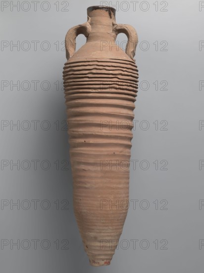 Amphora, unknown Egyptian workshop; amphorae, Egyptian amphorae, Egyptian ceramics, storage vessels, Coptic vessels, amphorae, amphorae, Nile clay. Wine transport vessel with a spindle-cylindrical, long body. Steep arms, cylindrical neck ending with a straight rim. Cylindrical vises, oval in cross-section, are attached to the lower part of the arms and to the middle part of the neck. The entire body has grooves of varying density and size; the densest are in the lower and upper parts. No cylindrical foot. Brown alluvial clay Naqlun AC1A ceramic type. Outer surface brown and even. The interior and the upper outer surface of the neck are coated with a black resinous substance. 2nd half of the 4th–5th century, Edfu find location. Vessel, amphora; height 65 cm, rim diameter 7 cm, body diameter 19 cm., A tall, narrow ceramic vessel with a rounded shoulder and a tapered base, featuring a pair of curved handles near the neck; the surface shows a series of horizontal ridges and grooves wrappin...