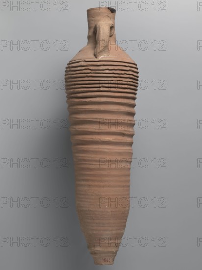 Amphora, unknown Egyptian workshop; amphorae, Egyptian amphorae, Egyptian ceramics, storage vessel, Coptic vessel, amphora; Nile clay. Wine transport vessel with a spindle-cylindrical, long body. Steep shoulders, cylindrical neck ending in a straight rim. Cylindrical vises, oval in cross-section, are attached to the lower part of the shoulders and to the middle part of the neck. Entire body has grooves of varying density and size; the densest are in the lower and upper parts. No cylindrical foot. Brown, alluvial clay Naqlun AC1A ceramic type. Outer surface brown and even. Interior and the upper outer surface of the neck coated with a black resinous substance. Dated 2nd half of the 4th–5th century. Findspot: Edfu. Vessel amphora. Height 65 cm; rim diameter 7 cm; body diameter 19 cm., A tall, elongated ceramic vessel with a narrow neck and a single handle shows a ribbed surface of uneven horizontal ridges extending along its body; the vessel is primarily terracotta brown with subtle vari...