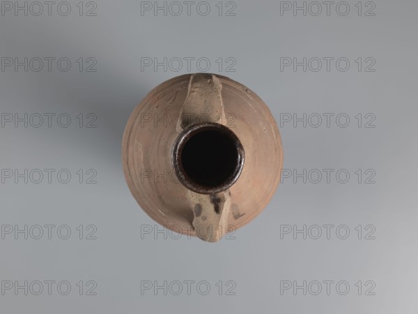 Amphora, unknown Egyptian workshop; amphorae, Egyptian amphorae, Egyptian ceramics, storage vessel, Coptic vessel, amphora; Nile clay. Wine transport vessel with a spindle-cylindrical, long body; steep arms; cylindrical neck ending in a straight rim. Cylindrical handles, oval in cross-section, are attached to the lower part of the arms and to the middle part of the neck. The entire body has grooves of varying density and size, densest in the lower and upper parts. No cylindrical foot. Brown alluvial clay Naqlun AC1A ceramic type. Outer surface brown and even; interior and upper outer neck coated with a black resinous substance. Dated 2nd half of 4th–5th century; findspot Edfu. Vessel amphora. Height 65 cm; rim diameter 7 cm; body diameter 19 cm., A top-down view of a round clay vessel with a dark circular opening at its center and a narrow handle crossing over the opening; the vessel is earthy brown with subtle variations and small darker spots, the handle appears a lighter beige, and ...