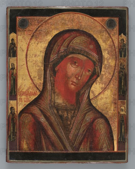 Icon: Our Lady of the Fiery One Ogniewidnaja, Old Believers' workshop, ca. 1850, Wietka; icon; height 35.5 cm, width 27.6 cm, A painted panel showing a close-up of a female figure with a tilted head, wearing a hooded garment in deep red with darker and lighter red shading and greenish brushstrokes, decorated with small star-like motifs and patterned trim; the face and neck are painted in reddish flesh tones with subtle shading and a somber expression, and the eyes look slightly downward. Behind the figure is a large circular halo rendered in gold with a textured, aged surface, set against a mostly gold background with areas of wear and darker mottling. Narrow vertical bands along the left and right edges contain small standing figures in darker colors including black, dark brown, and muted green, and there are dark circular medallions near the top corners. The outer border of the panel is dark, showing black and deep red tones, and the overall surface displays visible craquelure and si...