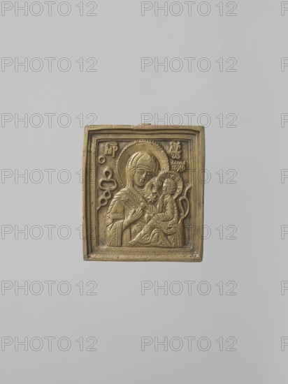 Icon: Our Lady of Tikhvin. Old Believers workshop. Our Lady of Tikhvin iconography; Mary mother of Jesus Christ; Jesus Christ Bible; icons; ornaments; Our Lady and Child iconography; maphorion iconography; inscriptions; unknown provenance. The icon represents a canonical variant of the Tikhvin image. The depiction of the figures, especially their faces, is somewhat archaizing. In the background of the image of the Mother of God with the Savior on her left shoulder, a schematic geometric pattern is marked. Inscriptions: MP OY, IC XC. On the reverse — a stylized floral-geometric ornament. 18th century, Russia. Icon. Height 5.8 cm; width 5.2 cm., A small rectangular relief panel in a metallic gold color is centered against a plain light gray background; the panel shows a raised depiction of two robed figures with halos, one larger holding the smaller on their lap, with detailed folds in the garments and ornamental borders around the scene, and there are a few decorative motifs carved into...