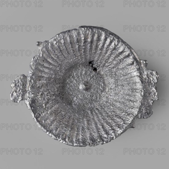 Miniature patera, repossession Przemysl, scenographic gallery class, model patera, votive offering, 1st–2nd century, Roman Empire 27 BC–476, toy/amulet; height 1 cm, width 3.7 cm, depth 4.2 cm, weight 0.014 kg, A roughly circular metallic object with a textured, cratered surface sits against a flat gray background; the object displays a silvery gray sheen with darker gray areas and subtle variations in tone, a concave central depression with radial ridges extending outward, irregular edges with a few protrusions and rough, pitted surfaces, and small dark voids near the center.
