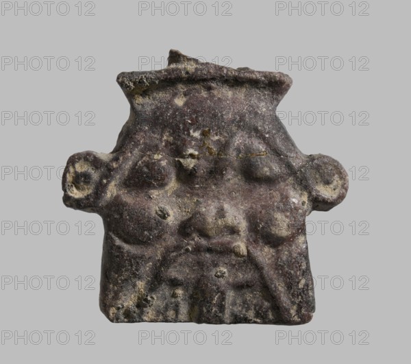 Plaque with a schematic relief representation of the head of Bes. The crown has been knocked off, protruding ears, the beard ends in a straight line. Amulet plaque, Late Period, Egypt, 7th–4th century BC. Height 2.7 cm; width 2.9 cm., A roughly square, weathered stone or clay object with an irregular top edge and small protrusions on the sides, showing a worn carved face-like relief with raised rounded features that resemble eyes and cheeks, a broad flat nose area, and a suggestion of a mouth and chin; the surface is textured with pits and erosion, showing a mix of dark brown, gray, beige, and muted yellowish spots against a neutral light gray background.