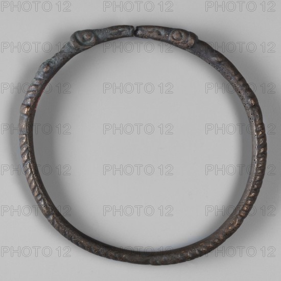 Bracelet with ram's head endings, unknown Roman workshop, repossession Silesia, bracelets, rams, 1st–3rd century, Roman Empire 27 BC–476, jewelry, weight 34 g, height 0.9 cm, diameter 7.5 cm, A circular metal ring with a textured surface lies flat against a light gray background, showing a roughly even round shape with a small gap at the top where the ends nearly meet. The ring's surface shows a mottled dark brown and black patina with areas of greenish-blue corrosion and occasional spots of brighter coppery brown. Raised ridges and a pattern of shallow grooves run along portions of the band, and the ends have slightly thicker, rounded terminal shapes with small raised bumps. The overall appearance is aged and weathered with uneven surface pitting and color variations in dark brown, black, greenish-blue, and coppery brown.