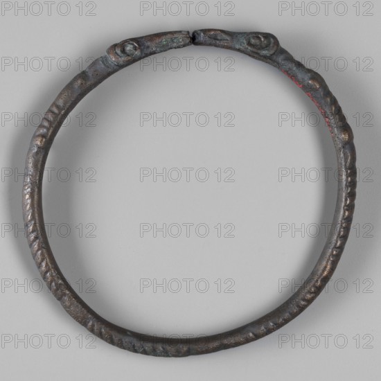 Bracelet with ram's head endings, unknown Roman workshop, repossession Silesia, bracelets, rams, 1st–3rd century, Roman Empire 27 BC–476, jewelry, weight 34 g, height 0.9 cm, diameter 7.5 cm, A circular metal bracelet with a textured surface and visible wear, showing dark brown and black tones with areas of greenish patina and small touches of red, set against a light gray background.