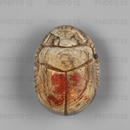 Scarab - a forgery? Scarab with a pseudo-inscription on the underside of the base. Traces of red dye visible on the surface of the wings. Probably a forgery. Contemporary product, Egypt. Scarab amulet. Height 1.2 cm; width 2.5 cm., An oval carved object resembling an insect form sits against a gray background; the object has a weathered, ivory-beige surface with darker brown hairline cracks and carved grooves defining segmented shapes, and several areas of worn red pigment on the lower central surface, with subtle variations of tan, cream, brown, and faded red across its textured surface.