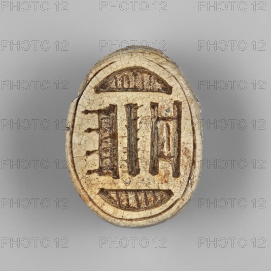 Scarab — possibly a forgery. Scarab amulet with a pseudo-inscription on the underside of the base. Traces of red dye on the surface of the wings. Probably a forgery; contemporary product, Egypt. Height 1.2 cm; width 2.5 cm., An oval, carved object in beige and brown tones sits against a light gray background; the surface shows a central recessed design made of darker brown incisions with symmetrical vertical and curved elements, and the beige surface has a network of fine cracks and a few darker brown spots and stains along the edges.