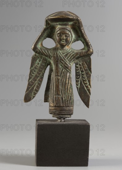 Figurine of a winged woman. Figurine depicting a woman standing frontally. Round face with clearly defined eyebrows, oval eyes, small slightly parted mouth, and rounded cheeks. Face framed by a headdress resembling a scarf. Raised hands hold a vessel in the shape of a bowl or basket placed on the head. Wings with schematically marked feathers descend vertically from both shoulders. Dressed in a robe with clearly defined vertical folds of drapery; hemline with horizontal stripes. A sash decorated with cross-shaped signs runs across the torso and arm. Figurine, height 4.5 cm, width 4.3 cm, weight 0.092 kg.,  unspecified date., A small metal figurine with a greenish-brown patina stands on a dark rectangular base; the figure has a rounded head with a simple facial expression, arms raised to hold a flat object above its head, and a long draped garment with incised vertical folds. The figure also has wing-like extensions on either side decorated with carved feather-like patterns, and areas o...