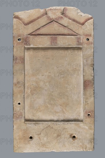 Stela Alexis, Small limestone stele with simple architectural decoration. Recessed epigraphic field surrounded by a wide strip; simple pediment and acroterions. Remains of engraved guide lines made by a stonemason. Traces of red paint highlighting elements of the pediment and on the top and side strips, in metopic form. Below the epigraphic field: an unidentified brownish, rounded shape. Side edges rather carelessly worked; back surface with very long tool marks. Object glued from eight fragments; upper part not preserved. Six later holes for wall mounting photos show the stele mounted to a wall. Dated 30 BC–1 AD beginning of the Roman period. Probable findspot: Tell el-Yahudiya. Grave inscription in Greek. Height 48.8 cm, width 27.4 cm, depth 2.7 cm., The image shows a rectangular stone tablet with a triangular pediment at the top and a recessed central panel; the stone surface is mostly beige with areas of pale tan and light brown, with scattered patches of reddish discoloration and ...