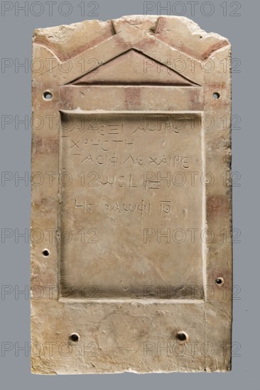Small limestone stele with simple architectural decoration. Recessed epigraphic field surrounded by a wide strip. Simple pediment and acroteria. Remains of engraved guide lines made by a stonemason while designing the decoration. Traces of red paint highlighting elements of the pediment and on the top and side strips in the form of metopic decoration. Below the epigraphic field, an unidentified brownish rounded shape. Side edges rather carelessly worked; back surface with very long tool marks. Object glued together from eight fragments; upper part not preserved. Six holes for mounting on the wall, probably added later for exhibition purposes. Greek funerary inscription. Date: 30 BC–1 AD beginning of the Roman period. Possible findspot: Tell el-Yahudiya. Height 48.8 cm, width 27.4 cm, depth 2.7 cm. Maker: unknown., A carved rectangular stone plaque with a raised border and a triangular pediment at the top, showing a weathered surface with areas of pale beige, cream, light brown, and fai...