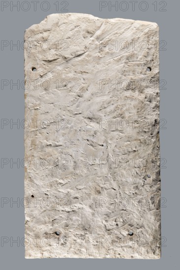 Small limestone stele with simple architectural decoration. Recessed epigraphic field surrounded by a wide strip. Simple pediment and acroteria. Remains of engraved guide lines made by a stonemason. Traces of red paint highlighting elements of the pediment and on the top and side strips in a metopic pattern. Below the epigraphic field a brownish rounded shape, unidentified. Side edges carelessly worked; back surface with very long tool marks. Object glued from eight fragments; upper part not preserved. Six holes for wall mounting, probably added later for display photographs show the stele mounted this way. Greek funerary inscription. Date: 30 BC–1 AD beginning of the Roman period. Possible findspot: Tell el-Yahudiya. Height 48.8 cm, width 27.4 cm, depth 2.7 cm. Maker: unknown., A textured rectangular slab with rough, uneven surfaces and visible tool marks, showing a mix of light colors including off-white, beige, and pale gray, with a few small darker brown and tan patches; the slab h...