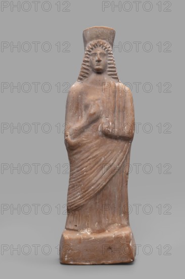 Figurine of a woman with a polos on her head, unknown Boeotian workshop, female figurine, 475–450 BC, Boeotia; sculpture; height 26 cm, width 9 cm, depth 7 cm, weight 0.43 kg., A standing terracotta figure of a person wearing a tall headdress and a long draped garment, shown from front-view on a rectangular base; the figure has stylized wavy hair framing the face, a simplified facial expression, one arm bent across the torso with the hand near the chest and the other arm bent with the hand resting near the waist, and the drapery is indicated by carved diagonal and vertical folds. The visible colors are shades of brown and tan on the figure with lighter worn spots and small areas of white residue, set against a neutral gray background.