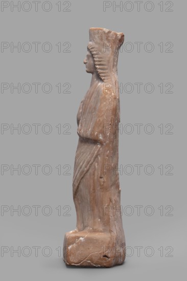 Figurine of a woman with a polos on her head, unknown Boeotian workshop; female figurine, 475–450 BC, Boeotia; sculpture; height 26 cm, width 9 cm, depth 7 cm, weight 0.43 kg., A side view of a standing carved figure in a warm brown stone with lighter veins and worn white patches, showing a profile face with a prominent nose and stylized hair or headdress that forms a ridged vertical band down the back; the figure wears a long draped garment with carved folds and holds its arms close to the body, all mounted on a rectangular base of the same brown stone, set against a plain neutral gray background.
