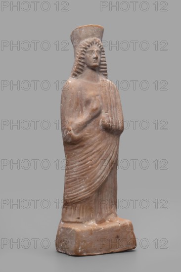 Figurine of a woman with a polos on her head, unknown Boeotian workshop, female figurine, 475–450 BC, Boeotia, sculpture; height 26 cm, width 9 cm, depth 7 cm, weight 0.43 kg., The image shows a standing terracotta statue of a robed figure against a plain gray background. The figure has a tall, rounded headdress and stylized, ridged hair framing the face, with a calm, neutral expression. The body is wrapped in draped garments with visible pleated folds and a hand positioned near the chest. The sculpture rests on a rectangular base with worn edges and surface abrasions. The visible colors are muted terracotta brown on the statue and base, lighter beige highlights on worn areas, and a neutral gray background.