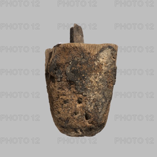 Beard from a sarcophagus mask, sarcophagus fragments, unknown provenance; beards facial hair, masks. Short beard tapering to a gently rounded bottom. Flat upper surface, with preserved mounting pin. Black paint preserved in places. ca. 1550–332 BC; New Kingdom–Late Period, Egypt. Possibly from a sarcophagus mask or figurine. Height 6.5 cm, width 4 cm., The image shows a single irregularly shaped object with a rough, weathered surface suspended against a plain light gray background; the object has an overall brown tone with darker brown and blackened patches, areas of lighter beige and tan, and hints of gray; its texture appears fibrous and pitted with a few small holes and indentations, and a narrow protrusion extends from the top.