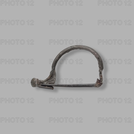 Aucissa-type fibula, Rome Empire, end of the 1st century BC–1st century. Complete brooch with a flat ring decorated with an elongated roller; earth-brown patina. Half of the thin roller, with a hole for attaching a needle, is broken off. Black Sea region origin; fibula; height 3.2 cm, width 5 cm, depth 1.4 cm, weight 6 g., An object resembling a curved metal pin or clasp sits against a plain light gray background; the metal shows a weathered, dark gray surface with areas of brownish rust and hints of greenish patina, a rounded knob at one end, a slender straight rod spanning the lower edge, and a semicircular arch forming the upper part, with rough, irregular textures and slight shadowing beneath the object.