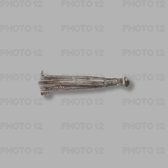 Aucissa-type fibula, Rome Empire. Complete brooch of the Aucissa type from the Black Sea region. Flat ring decorated with an elongated roller; earth-brown patina. Half of the thin roller, with a hole for attaching a needle, is broken off. End of the 1st century BC–1st century. Unknown site of origin Black Sea region. Fibula; height 3.2 cm, width 5 cm, depth 1.4 cm, weight 6 g., A single elongated metallic object lies horizontally against a plain light gray background, showing a weathered surface with patina in shades of dark gray, brown, and hints of green; one end tapers into a rounded, bulbous knob while the opposite end is slightly flared with a small protrusion, and the central shaft displays a series of longitudinal ridges and shallow grooves giving it a segmented appearance.
