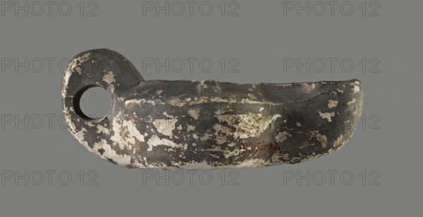Oil lamp, unknown Gaulish workshop role/classification of maker or workshop uncertain, from 2nd century Gaul. Lamp from a mold. Round vessel with arms sloping outward, separated from the disc by a convex edge. Flat, concave disc with an almost centrally located oil hole. Two bars placed symmetrically on the arms; third vice placed vertically opposite the burner. Long wick channel decorated in the center with a wide flat groove bounded by two convex edges. Rounded end of the burner; large wick hole; no charring. Traces of a faded trademark on the bottom. Type: Buchi type Xa Loeschcke type X oil lamp. Dimensions: height 3.3 cm, width 11.8 cm, depth 6.6 cm., An object resembling a shallow bowl with a rounded handle and a circular hole is shown against a plain background; the surface is mottled with dark gray and black areas and worn patches of off-white and beige, with irregular flaking and rough texture visible across the form.