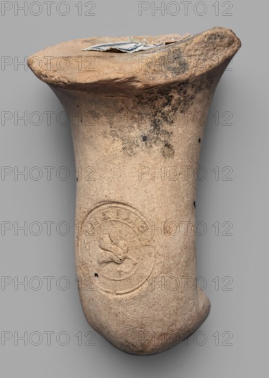 Fragment of a transport amphora neck stamped with a Rose of Rhodes, unknown Rhodian workshop, stamp impressions, trade. Fragment of a transport amphora neck stamped with a Rose of Rhodes and the inscription Chresimou, ca. 280–ca. 220 BC, Rhodes; vessel stamped neck, height 6.8 cm, width 10 cm., A single clay object with a flared rim and tapered body rests against a neutral gray background; the surface is rough and weathered with a base color of light tan and areas of darker brown and gray discoloration, including some blackish spots and smudges; the rim shows irregular edges and a few pieces of torn paper or cloth in pale white and blue sitting on top; the body bears a circular impressed design with raised relief motifs and surrounding rings, and the lower end of the object is rounded and slightly darker from wear.