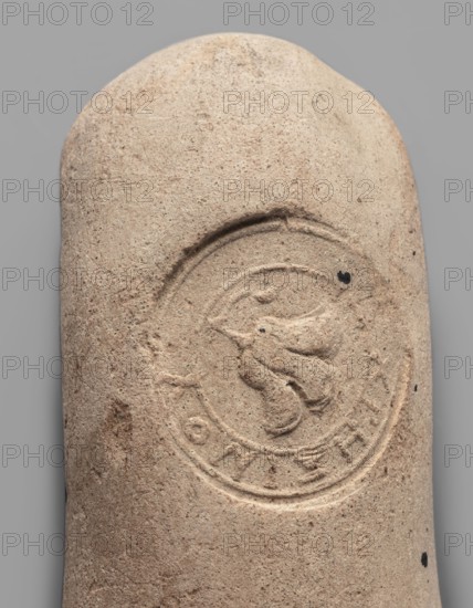 Fragment of a transport amphora vice stamped with a Rose of Rhodes and the inscription Chresimou, unknown Rhodian workshop, ca. 280–ca. 220 BC, Rhodes; vessel stamped vice, height 6.8 cm, width 10 cm., A close-up view of a rounded, cylindrical object with a rough, grainy surface in beige and light tan tones, showing an incised circular emblem with raised and recessed carved shapes in the center and a few small dark spots on the surface; the background is a uniform medium gray.