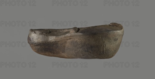 Oil lamp, unknown Germanic workshop or other provincial?. Molded lamp. Round vessel. Arms sloping outward, separated from the disc by a convex edge. Three bars arranged on the arms two symmetrically on the sides, the third opposite the burner. Concave, flat disc with one large centrally located oil hole. Medium-length wick channel, decorated in the center with a wide flat groove bounded by two convex edges. Rounded end of the burner. Large wick hole. No blackening. 1st–2nd century AD, Roman Empire 27 BC–476. Oil lamp. Height 3.4 cm; width 8 cm; depth 5.3 cm. Buchi type Xa = Loeschcke type X lamp., A single elongated, irregularly shaped object with a rounded bottom and a slightly concave top floats against a plain gray background; its surface shows a rough, uneven texture with pits and small bumps, and the visible colors include dark brown, muted brown, gray, and hints of black and olive tones, with some lighter brown highlights along raised areas.