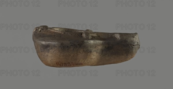 Oil lamp, unknown Germanic workshop or other provincial?, molded round vessel with arms sloping outward separated from the disc by a convex edge. Three bars on the arms two symmetrically on the sides, the third opposite the burner. Concave, flat disc with one large central oil hole. Medium-length wick channel decorated in the center with a wide flat groove bounded by two convex edges. Rounded end of the burner. Large wick hole. No blackening. 1st–2nd century AD, Roman Empire 27 BC–476. Oil lamp; height 3.4 cm, width 8 cm, depth 5.3 cm. Type: single-burner Roman lamp, Buchi type Xa / Loeschcke type X., A roughly boat-shaped object with an uneven, worn surface in shades of brown, dark brown, and gray sits against a gray background; the object has rounded edges, a shallow hollow along its top, a few small dents and cracks, and areas of lighter brown and darker brown mottling giving a textured, weathered appearance.