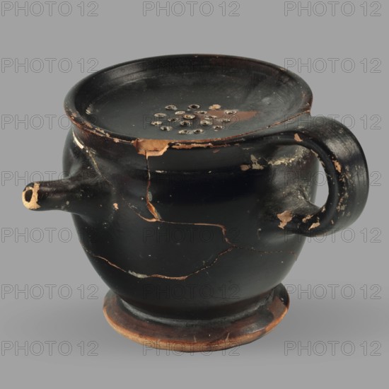 Askos with spout and strainer, unknown Attic workshop. Greek pottery toilet vessel with tubular spout and ribbon handle; top covered with a strainer. 5th century BC, Attica. Vessel askos. Height 6.2 cm, width 8.3 cm, depth 9 cm, weight 0.09 kg., A rounded ceramic vessel with a wide mouth and a raised foot, shown against a plain gray background; the vessel is primarily black with exposed areas of brownish-orange where the surface is chipped or worn, a few lighter beige patches around damaged edges, and several thin hairline cracks revealing brown beneath; it has a curved handle on one side and a short spout on the opposite side, and the top surface features a cluster of small circular indentations; the base rim shows visible wear in brown and the overall surface has a matte black finish with scattered areas of chipped brown-orange.