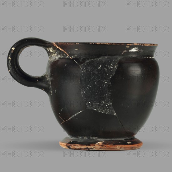 Askos with spout and strainer, unknown Attic workshop, Greek pottery toilet vessel with tubular spout and ribbon handle; top covered with a strainer. 5th century BC, Attica; vessel askos. Height 6.2 cm, width 8.3 cm, depth 9 cm, weight 0.09 kg., A single-handled cup with a rounded body and a flared rim sits against a plain gray background; the cup is primarily dark brown to black with worn areas revealing terracotta and lighter brown at the rim, handle edge, and foot, and there are visible repairs where fragments meet, showing lighter lines and patches, as well as scattered small pale spots and scuffs across the surface.