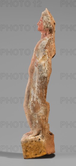 Male figurine, unknown Boeotian workshop. Standing man on a high quadrangular base, arched at the back. The weight of the body rests on the right leg, the left leg slightly forward. Torso naked at the front, leaning backward with the right hip protruding; slender body. A robe draped over the shoulders and covering the back conceals the hands. Head slightly tilted to the right; face treated schematically with large eyes. In the lowered right hand the figure holds an elongated object, while the left hand presses a round vessel to the chest. The base is covered with a visible strip of brown paint; the figurine bears traces of red-pink polychrome and the eyes were covered with white paint. 5th–4th century BC earlier dating: mid-5th century BC. Boeotia. Terracotta figurine, height 27 cm, width 6.8 cm, depth 6 cm, weight 0.31 kg., A weathered standing figurine shown in profile, carved from a clay-like material with a rough, pitted surface and worn edges; the figure has a simplified human for...