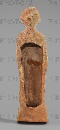 Male figurine, unknown Boeotian workshop. Standing man on a high quadrangular base, arched at the back. Weight of the body rests on the right leg, left leg slightly forward. Torso naked at the front, leaning backward with the right hip protruding; body slender. A robe draped over the shoulders and covering the back conceals the hands. Head slightly tilted to the right; face treated schematically with large eyes. In the lowered right hand the figure holds an elongated object, while the left hand presses a round vessel to the chest. Base covered with a visible strip of brown paint; figurine bears traces of red-pink polychrome. The eyes were covered with white paint. 5th–4th century BC earlier dating: mid-5th century BC. Boeotia. Terracotta figurine. Height 27 cm, width 6.8 cm, depth 6 cm, weight 0.31 kg., A standing human-shaped terracotta figure viewed from the front with a stylized rounded head and narrow shoulders, showing a hollowed-out torso cavity; the surface is primarily warm ora...