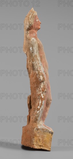 Male figurine, unknown Boeotian workshop. Standing man on a high quadrangular base, arched at the back. The weight of the body rests on the right leg, the left leg slightly forward. Torso naked at the front, leaning backward with the right hip protruding; slender body. A robe draped over the shoulders and covering the back conceals the hands. Head slightly tilted to the right; face treated schematically with large eyes. In the lowered right hand the figure holds an elongated object, while the left hand presses a round vessel to the chest. Base bears a visible strip of brown paint; the figurine shows traces of red-pink polychrome and the eyes were covered with white paint. 5th–4th century BC earlier dating: mid-5th century BC, Boeotia. Terracotta figurine; height 27 cm, width 6.8 cm, depth 6 cm, weight 0.31 kg., A weathered standing figurine viewed in profile on a rectangular base, rendered with an elongated torso and head looking forward; the surface shows worn patches of pinkish-red, ...